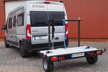 Der unbeladene SMV Alu-Star Rolli angekuppelt an einen Camper. Freie Sicht auf Merkmale des Anhängers wie Haltearm, Auffahrrampe, Sicherungsgurte und Keile.