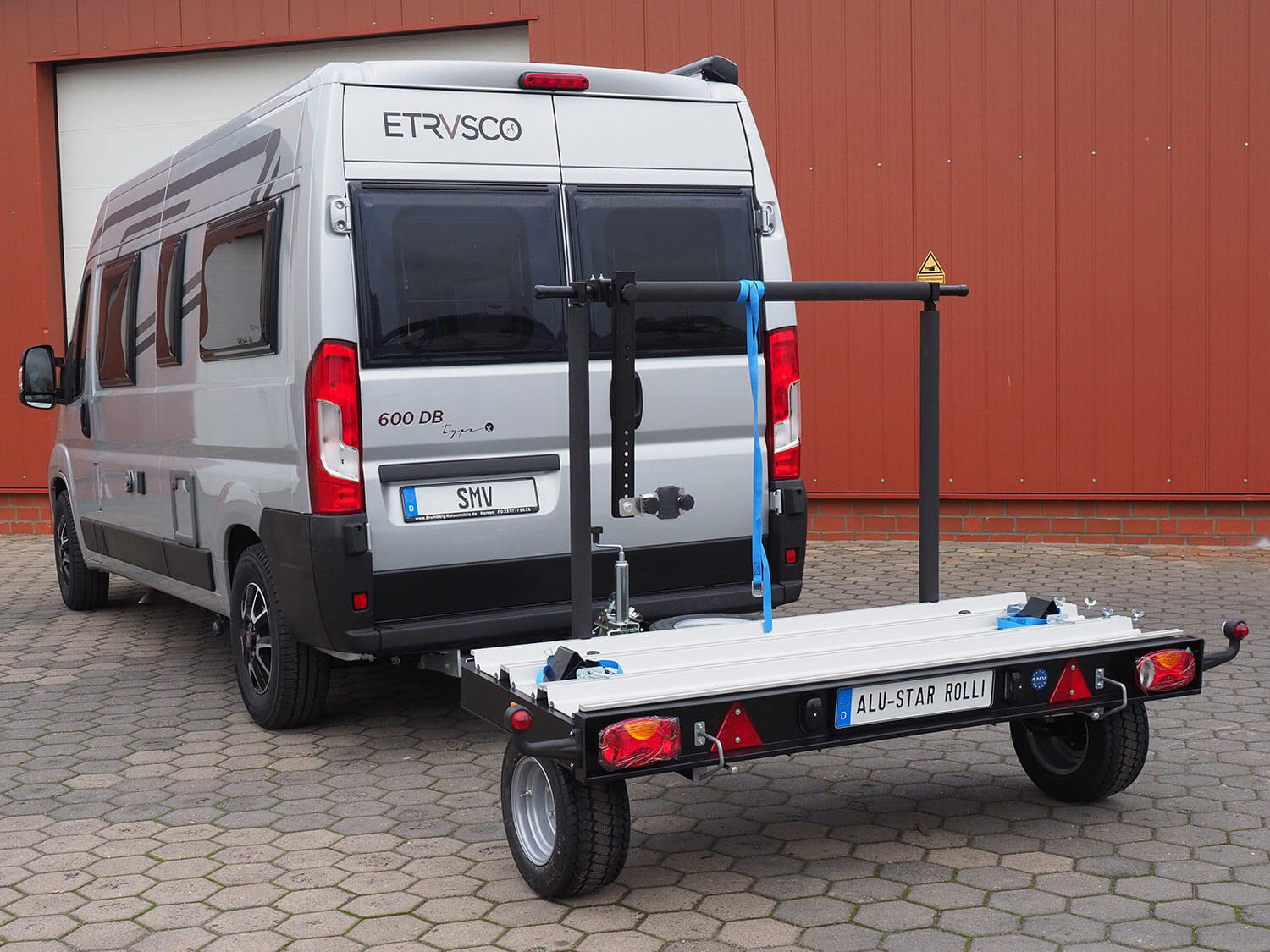 Der unbeladene SMV Alu-Star Rolli angekuppelt an einen Camper. Freie Sicht auf Merkmale des Anhängers wie Haltearm, Auffahrrampe, Sicherungsgurte und Keile.