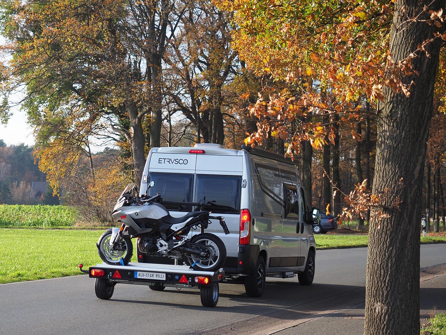 Ein Campingmobil mit dem Anhänger "Alu-Star Rolli" von SMV ist auf einer herbstlichen Baumallee vorbeigefahren. Auf dem Anhänger wird ein Motorrad transportiert.