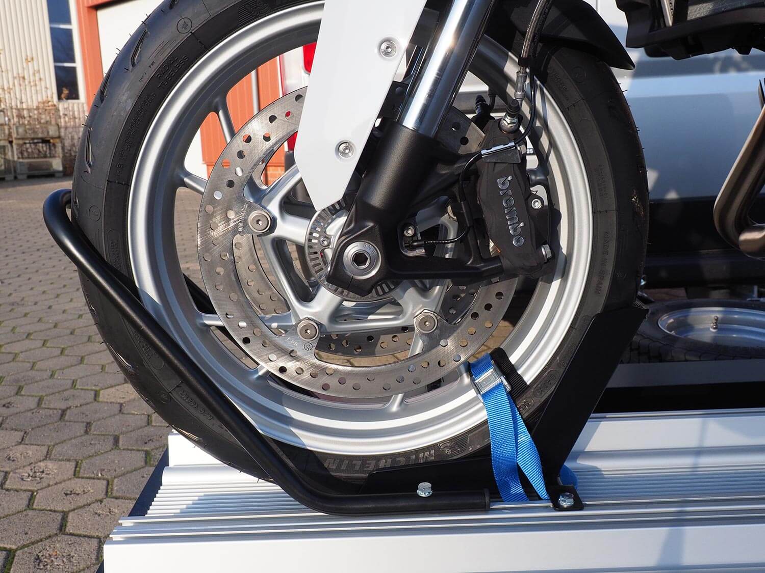 SMV Alu-Star Rolli. Das Vorderrad eines Motorrades gesichert mit Wippe, Reifenhaltebügel und Spanngurt auf der Plattform.