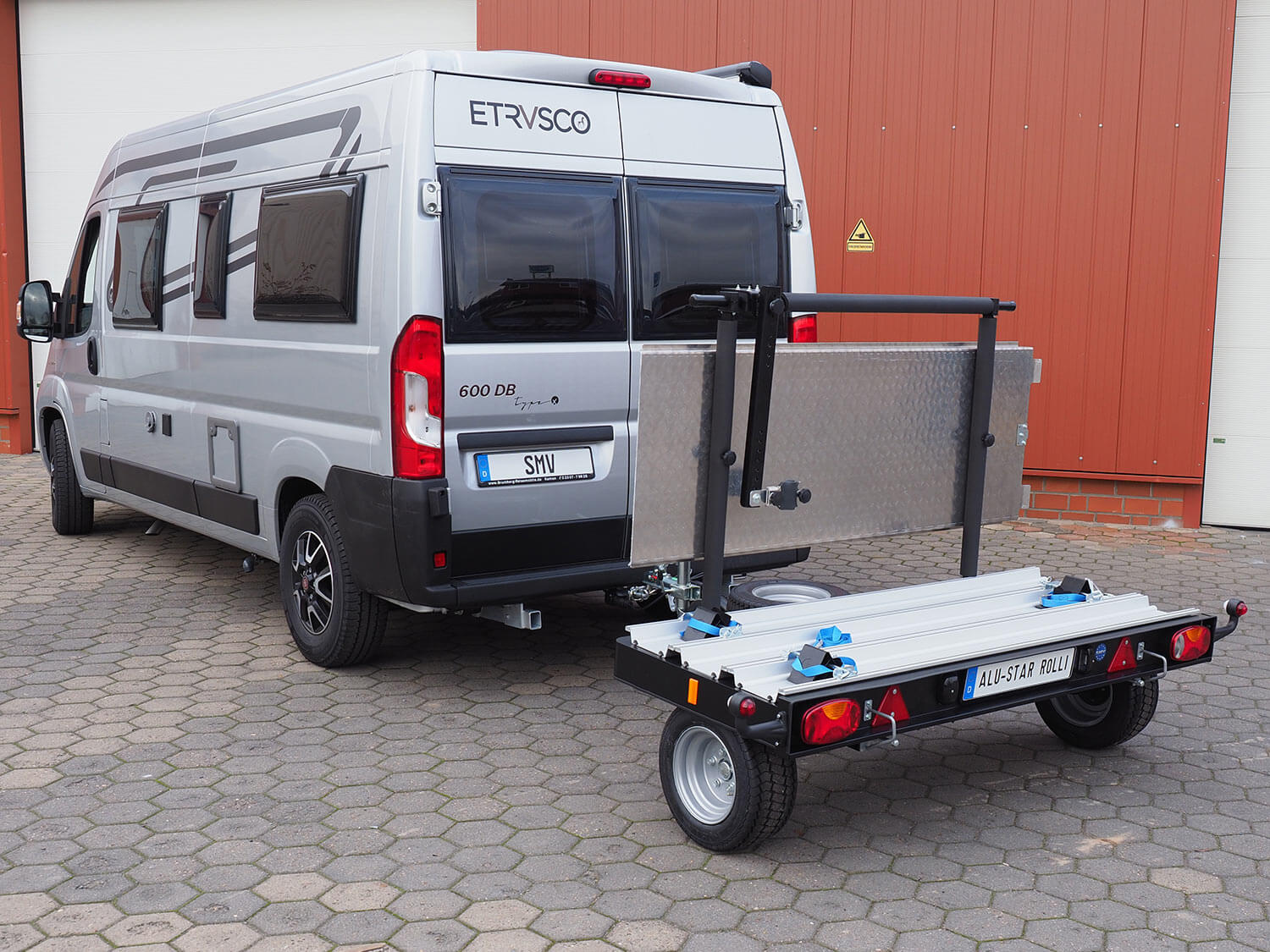 Der unbeladene SMV Alu-Star Rolli MP3 angekuppelt an einen Camper. Freie Sicht auf Merkmale des Anhängers wie Haltearm, Auffahrrampe, Sicherungsgurte und Keile.