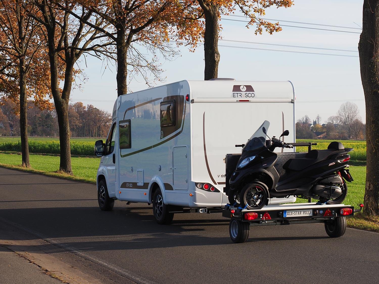SMV Alu-Star Rolli MP3. Ein Wohnmobil mit dem Anhänger "Alu-Star Rolli MP3" von SMV ist auf einer herbstlichen Baumallee vorbeigefahren. Auf dem Anhänger wird ein dreirädriger Roller transportiert.