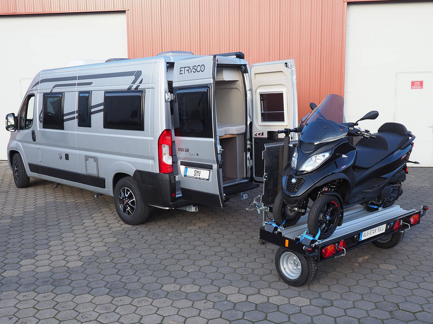 Der SMV Alu-Star Rolli MP3 steht mit einem MP3 Roller beladen hinter einem Camper mit geöffneten Hecktüren.