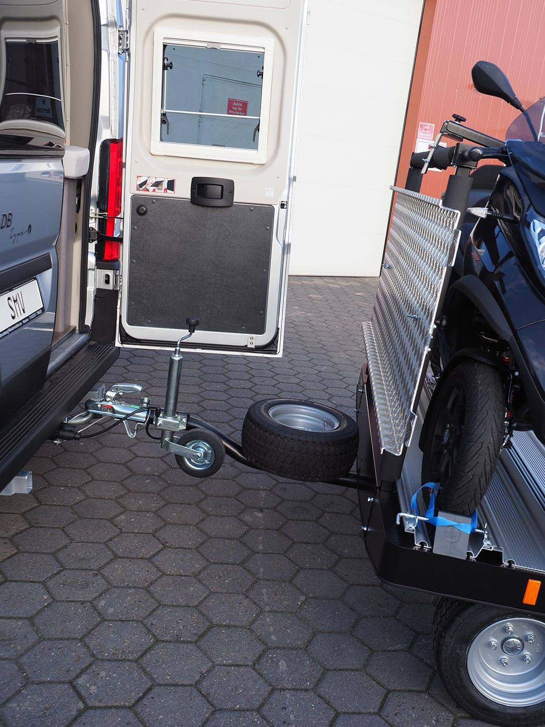 Der SMV Alu-Star Rolli MP3 ist an einen Camper angehängt und mit einem MP3 Roller beladen. Die rechte Hecktür ist geöffnet und es ist genügend Abstand bis zum Anhänger. Auf der Deichsel ist das Reserverad befestigt.