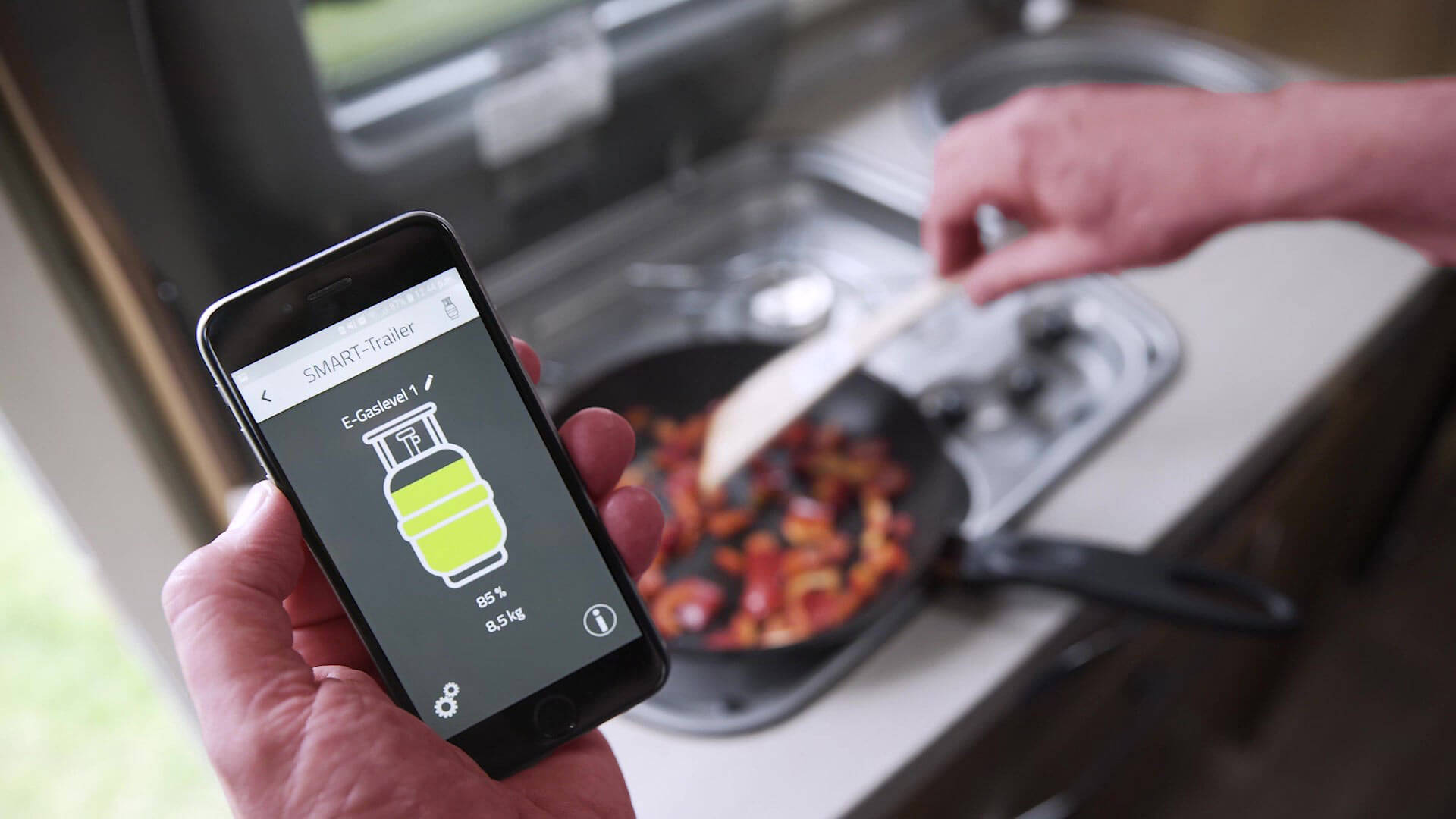 Eine Hand hält Smartphone auf dem in der App der Füllstand einer Gasflasche angezeigt wird, während die andere Hand im Hintergrund Essen zubereitet.