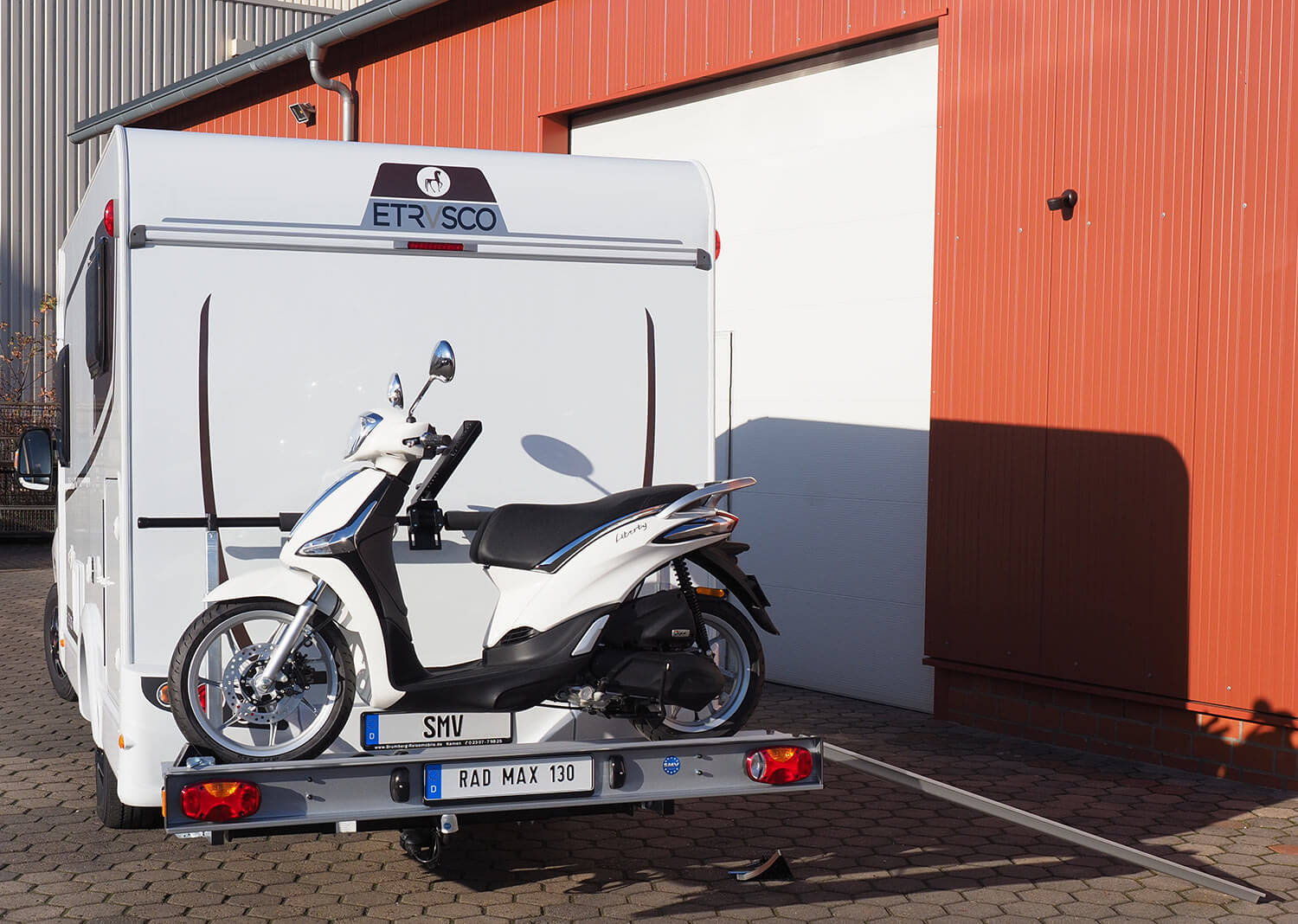 SMV Rad Max 130 mit einem Roller beladen und angebauter Auffahrrampe. Heckansicht eines Wohnmobils.